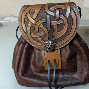 May include: A brown leather pouch with a flap featuring a Celtic knot design. The flap is secured with a brass button. The pouch has a drawstring closure and a black strap.