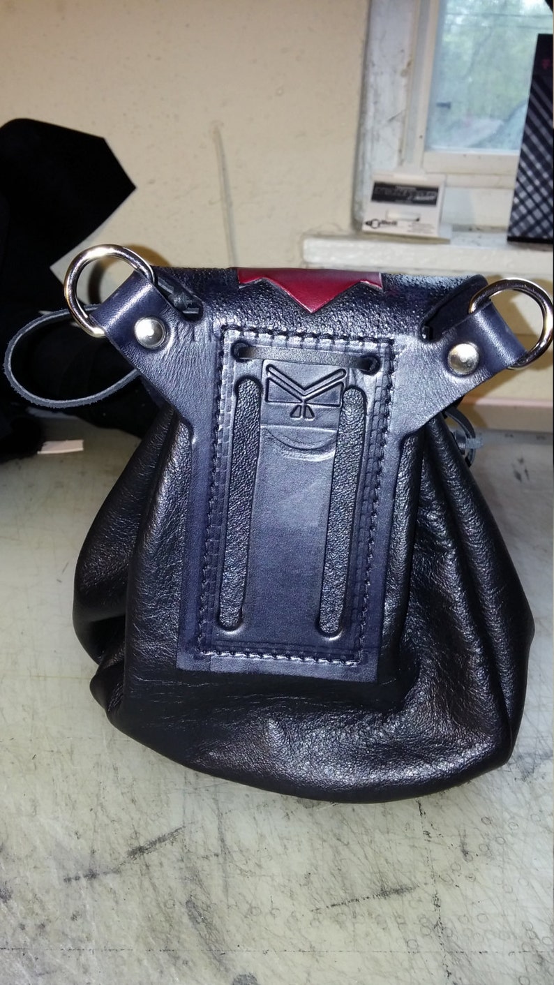 May include: Black leather pouch with a red and black flap. The pouch has a decorative stitched design and two metal rings.