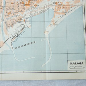 May include: A vintage map of Malaga, Spain, showing the city's port and surrounding areas. The map is printed on a light blue background with brown lines and text. The map includes a scale in meters and a legend in Spanish.