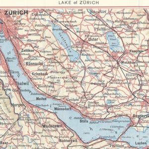 May include: A vintage-style printed map titled Lake of Zurich, featuring the blue water of the lake surrounded by tan land, red road lines, and black text labels for various towns and cities, with a scale bar at the bottom and a red border.