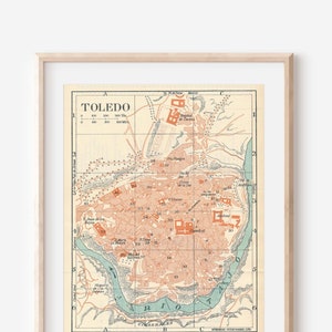 May include: A vintage map of Toledo, Spain, printed in shades of brown, green, and blue. The map shows the city's streets, buildings, and landmarks, including the Alcazar and the Cathedral. The map is framed in a light wood frame.