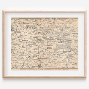 May include: A framed vintage map print. The map features detailed lines and text, with a beige and gray color scheme. The map is encased in a light brown wooden frame. The print is a historical representation of a geographical area.