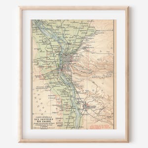 May include: A framed vintage map titled Carte Générale des Environs du Caire, featuring detailed topography, river paths, and geographical markings in muted green, tan, and brown tones, presented within a simple light-colored wooden frame.