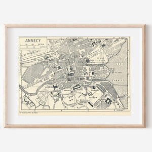 May include: A framed black and white map of Annecy, France, featuring detailed street layouts, building blocks, and the Lac d'Annecy. The map includes a grid system labelled with letters and numbers, a scale bar, and clear text labels for various city landmarks and districts.