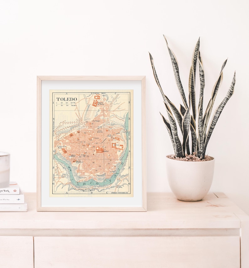 May include: A framed vintage map of Toledo, Spain, with a light brown frame and a white mat. The map is in shades of blue, green, and red, with the city's streets and landmarks labeled in black text. The title "TOLEDO" is printed in bold, black letters at the top of the map.