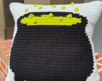 May include: A crocheted pillow with a black cauldron design and green bubbling potion. The pillow is white with a black border.