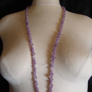 May include: A long necklace made of small, irregular amethyst beads. The beads are a light purple color and are strung together on a clear thread. The necklace is displayed on a white mannequin.