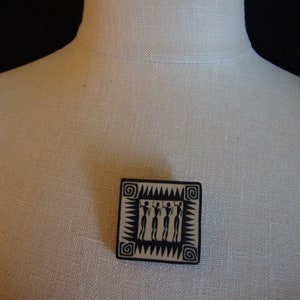 May include: A black and white ceramic brooch with a geometric design featuring four figures in a row. The figures are stylized and have a tribal feel.