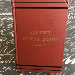May include: A vintage copy of "Roberts Rules of Order Revised" book. The book has a red cover with gold lettering and black stripes. The book is held by a gold-colored stand.