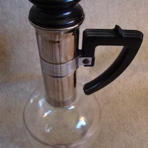 May include: A clear glass bottle with a silver and black top and a black handle. The bottle has a round body and a long neck.
