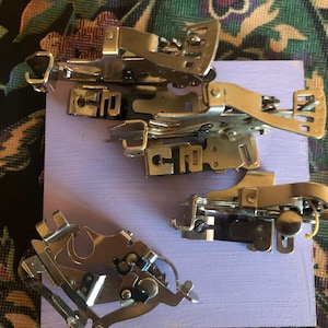 May include: A collection of silver sewing machine presser feet, arranged on a light purple wooden block. The feet have various shapes and designs, with levers and attachments. The image showcases sewing accessories.