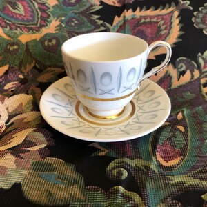 May include: A white teacup and saucer set with a light blue and gold design. The cup and saucer have a repeating pattern of geometric shapes. The set is on a patterned fabric background.
