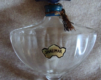 Könnte beinhalten: Eine durchsichtige Glasflasche mit einer strukturierten Oberfläche und einem goldenen Etikett mit der Aufschrift "Shalimar". Die Flasche hat einen blauen und grünen Verschluss mit dem Text "Guerlain Paris" darauf.