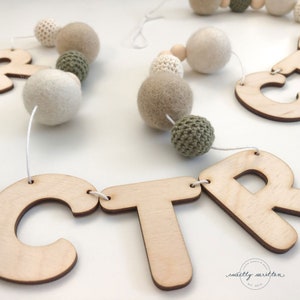May include: A wooden letter banner with the letters "CTR" strung together with white twine. The banner is decorated with beige and green felt balls and crocheted balls.