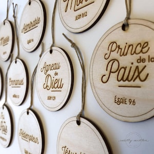 May include: Wooden circular ornaments with various scriptural phrases and verses, including "Prince de la Paix" and "Agneau de Dieu," are suspended by twine. The ornaments have a light wood finish with dark brown engraved text and borders.