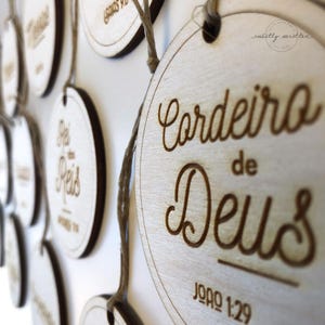 May include: Wooden, circular plaques with laser-etched text and a natural wood grain finish. The plaques are suspended by twine and feature religious phrases in Portuguese. The text "Cordeiro de Deus" is visible.