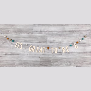 May include: A wooden banner with the words "It's Great To Be 8" strung with white, orange, and teal felt balls.