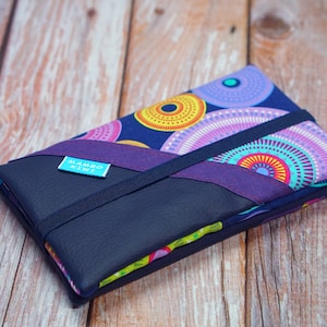 Può includere: Una custodia in tessuto blu navy e viola con un colorato motivo circolare. La custodia ha un elastico blu scuro e una piccola etichetta blu con la scritta "Mambo Kiwi". La custodia è chiusa e si trova su una superficie di legno.