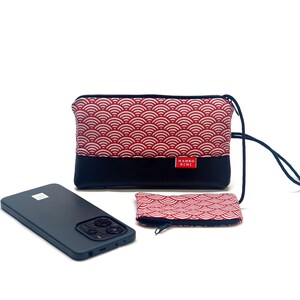 Puede incluir: Un smartphone negro junto a un conjunto de accesorios a juego. El conjunto incluye una bolsa grande con cremallera, una bolsa pequeña con cremallera y una correa para la muñeca. Las bolsas presentan un patrón de olas rojas y blancas en la mitad superior y una mitad inferior negra. La marca "MAMBO KIWI" está en la bolsa grande.