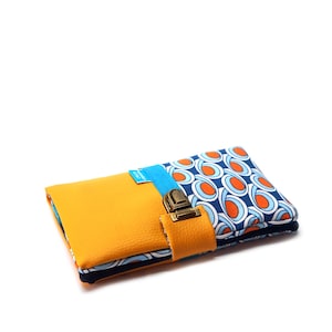 Puede incluir: Una cartera plegada con una parte delantera de cuero amarillo mostaza y una parte trasera de tela estampada. La tela presenta un diseño azul y blanco con detalles en naranja. Una correa azul y un cierre metálico aseguran la cartera.