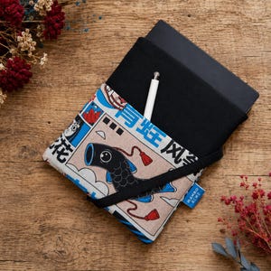 Puede incluir: Una funda de tela negra para tableta o cuaderno con un bolsillo frontal estampado que muestra un pez koi negro, detalles en azul y caracteres japoneses. Un lápiz óptico blanco está insertado en el bolsillo y una banda elástica negra asegura el estuche.