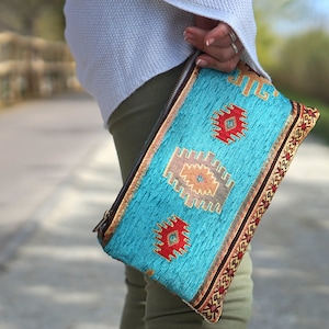 Könnte beinhalten: Eine rechteckige Clutch mit Reißverschluss, die eine leuchtend türkisfarbene Stoffbasis mit gewebten geometrischen Mustern in Rot, Braun und Creme aufweist. Die Tasche ist mit einer dekorativen Kante versehen, die ein sich wiederholendes rot-schwarzes Muster zeigt.