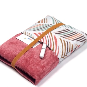 Può includere: Una custodia in tessuto rettangolare con una sezione inferiore in velluto a coste rosa polvere e una sezione superiore bianca con un motivo a foglie multicolore. Un sottile elastico marrone chiude la custodia, fissando una penna e una carta.