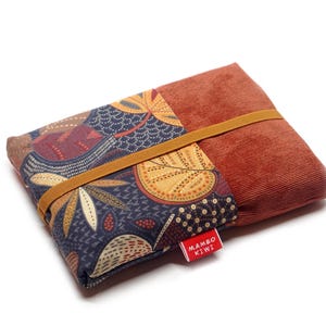 Puede incluir: Una bolsa rectangular de patchwork con textura de pana en tonos óxido y tela estampada con diseños de hojas y flores en azul marino, marrón y dorado. Una banda elástica marrón asegura la bolsa. Una etiqueta roja dice "MAMBO KIWI."