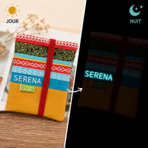 Könnte beinhalten: Eine rechteckige Stofftasche mit horizontalen Streifen in Rot, Schwarz, Türkis und Gelb, die mit einem roten Gummiband verschlossen ist. Der Name SERENA ist in Weiß aufgedruckt, leuchtet im Dunkeln und hat ein grünes Etikett MAMBO KIWI.