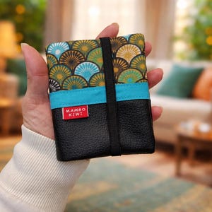 Peut inclure: Un petit portefeuille noir avec un motif de ventilateur coloré sur la moitié supérieure, une bande turquoise et une bande élastique noire. La moitié inférieure est en cuir noir. L'étiquette rouge indique "MAMBO KIWI".