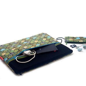 Może przedstawiać: Etui na laptopa i pasująca torba na akcesoria z kolorowym wzorem przypominającym łuski w odcieniach zieleni, niebieskiego i żółtego. Etui jest otwarte, ukazując czarny laptop i białą ładowarkę. Widoczna jest również mała torebka i akcesoria techniczne.