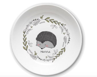Puede incluir: Plato de cerámica blanco con una ilustración de erizo gris, nariz rosa y una corona floral verde y gris. El nombre "Hanna" está escrito debajo. Un diseño encantador y caprichoso.