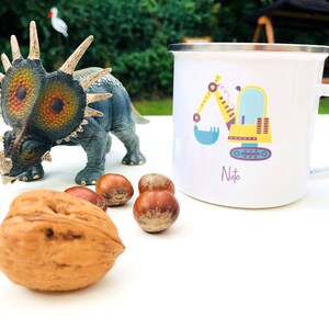 Könnte beinhalten: Weißer Emaillebecher mit einem farbenfrohen Bagger-Design und dem Namen "Nate" aufgedruckt. Ein blau-graues Dinosaurier-Spielzeug, Walnüsse und Haselnüsse sind ebenfalls im Bild. Der Becher hat einen silbernen Rand und einen weißen Henkel.