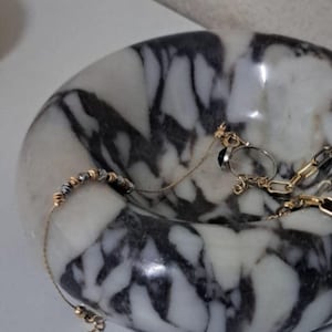 Könnte beinhalten: Eine weiße Marmor Schale mit schwarzen Adern, die als dekorative Schale für Schmuck verwendet wird. Die Schale ist gefüllt mit einer goldenen Kettenkette, einem goldenen Ring und einem silbernen Perlenarmband.