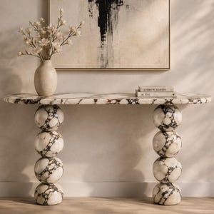 Puede incluir: Una consola de mármol con una tapa estampada y patas esféricas. La mesa sostiene un jarrón con flores blancas y una pila de libros. Una pintura abstracta en blanco y negro cuelga de la pared detrás de la mesa.