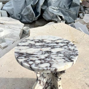 May include: A round, white marble table with a unique, three-legged base. The marble has a swirling pattern of dark purple and grey veins.