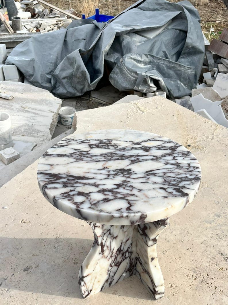 May include: A round, white marble table with a unique, three-legged base. The marble has a swirling pattern of dark purple and grey veins.