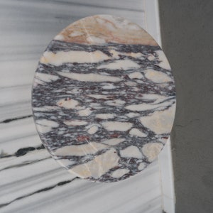 May include: A round, oval-shaped marble table top with a swirling pattern of white, grey, and pink.