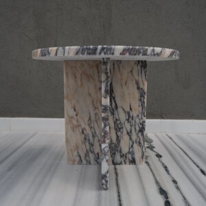May include: A round marble table with a white, pink, and purple marbled top and base. The base is made of four rectangular pieces of marble that intersect in the middle.