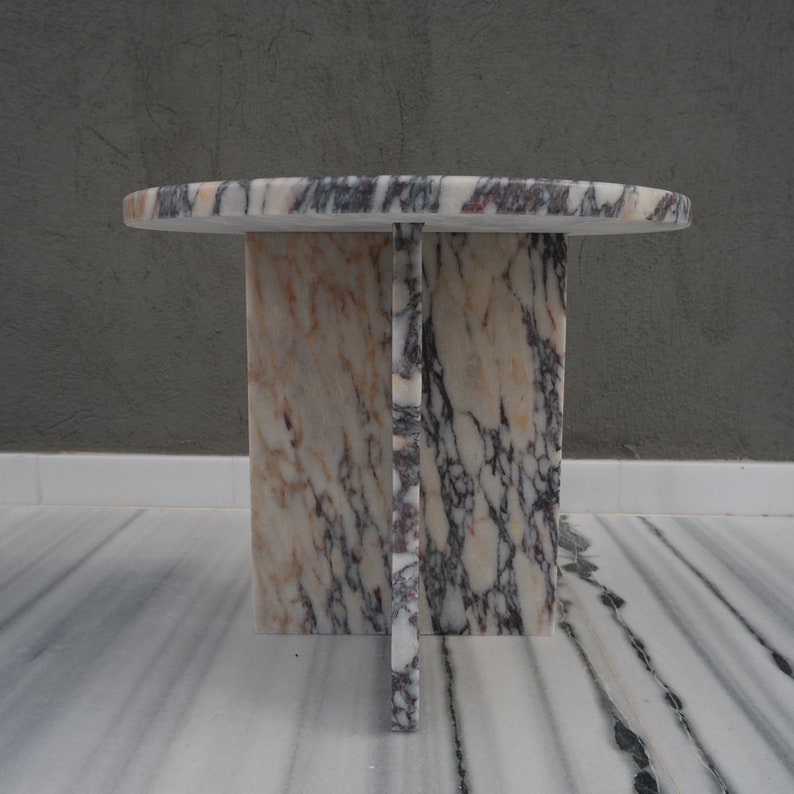 May include: A round marble table with a white, pink, and purple marbled top and base. The base is made of four rectangular pieces of marble that intersect in the middle.