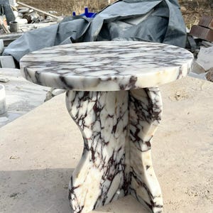 May include: A round, white marble table with a unique, abstract base. The table top has a swirling pattern of grey and purple veins.