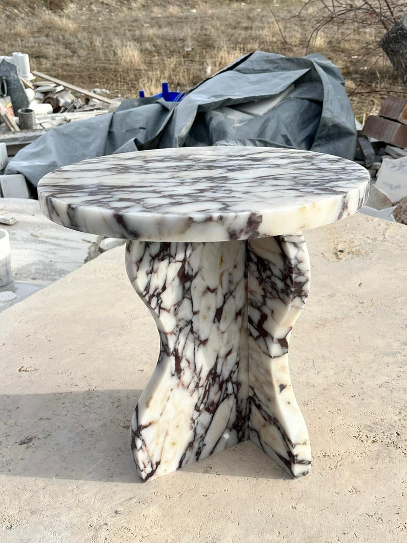 May include: A round, white marble table with a unique, abstract base. The table top has a swirling pattern of grey and purple veins.