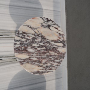 May include: A round, white marble coaster with a swirling pattern of pink, brown, and yellow veins. The coaster is on a white marble surface.