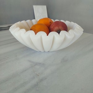 May include: White marble bowl with a fluted design, holding three pieces of fruit: two orange and one red.