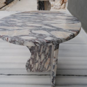 May include: A round, white marble table with a unique, swirling pattern of grey and purple veins. The table has a single, curved leg.