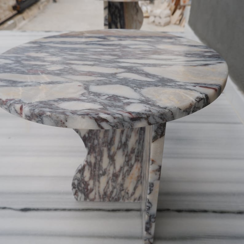 May include: A round, white marble table with a unique, swirling pattern of grey and purple veins. The table has a single, curved leg.