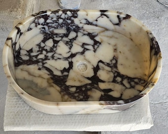 May include: Oval-shaped marble sink with a cream and dark brown marbled pattern. The sink has a drain hole and is set on a white surface. The sink's design is a natural stone pattern.