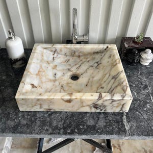 Könnte beinhalten: Ein quadratisches Waschbecken aus Marmor mit einem zentralen Abflussloch. Das Waschbecken hat ein cremefarbenes und braunes Adernmuster. Über dem Waschbecken ist ein verchromter Wasserhahn installiert. Ein Seifenspender und Dekorationsartikel befinden sich auf der Arbeitsplatte.
