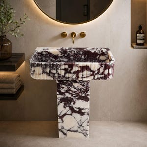 May include: A rectangular pedestal sink crafted from white and deep purple veined marble. The basin features a fluted exterior design with vertical grooves. It is paired with a gold-toned wall-mounted faucet fixture centred above the basin.