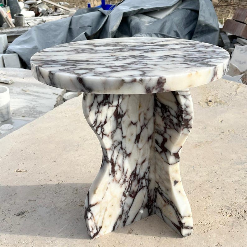 May include: A white marble side table with a round top and a unique, abstract base. The base is made of white marble with brown and grey veins.